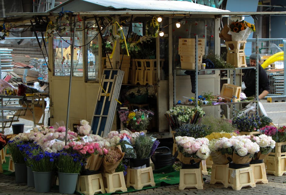 Image of a busy flower stall at Brixton Market featuring an array of fresh floral arrangements. The foreground displays numerous bouquets of flowers, including pink peonies, white roses, lavender, and