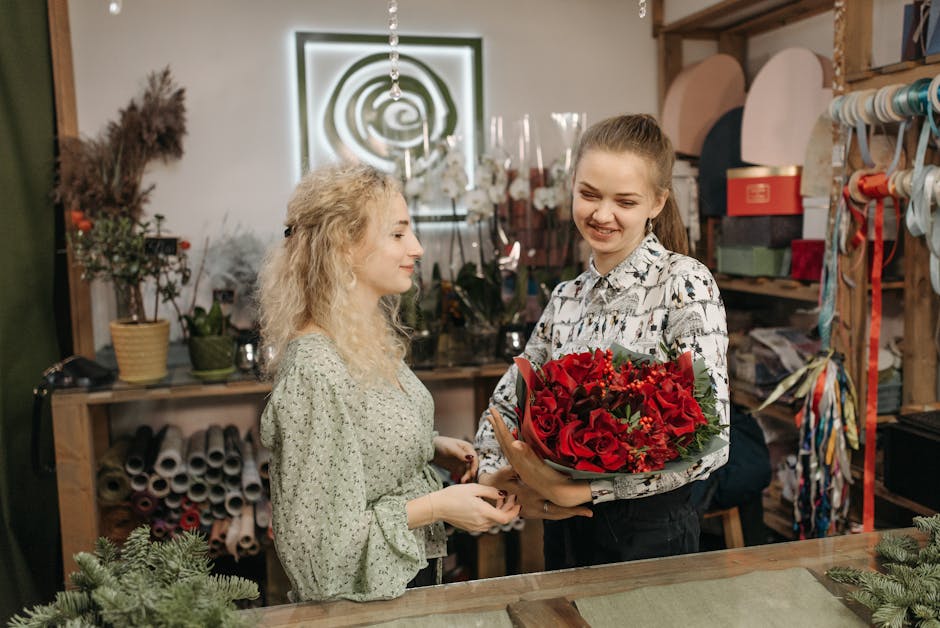 A vibrant display of floral arrangements and bouquets featuring red roses, white sprigs, and greenery, arranged in baskets and wrapping paper. Several heart-shaped red foil balloons are interspersed a