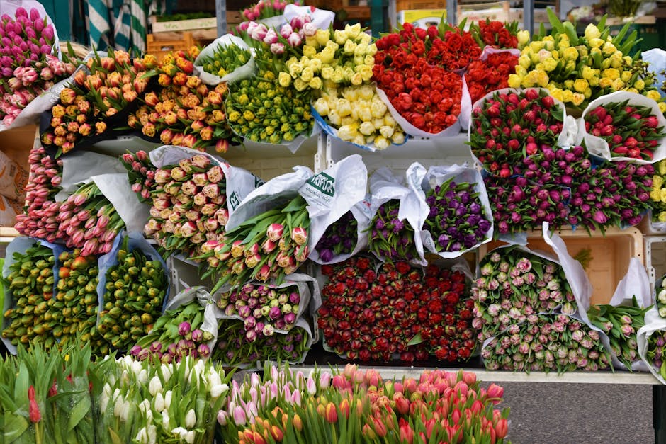 A vibrant display of freshly cut flowers arranged in bouquets and bundles at a florist market, featuring tulips in shades of pink, white, and red, along with rose varieties in yellow, red, purple, and