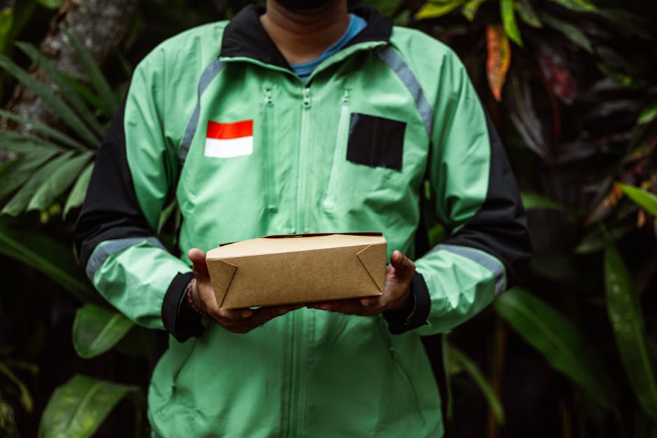A person wearing a green delivery jacket with a black patch and an Indonesian flag patch on the chest holds a rectangular cardboard parcel with both hands. The background features lush green foliage,