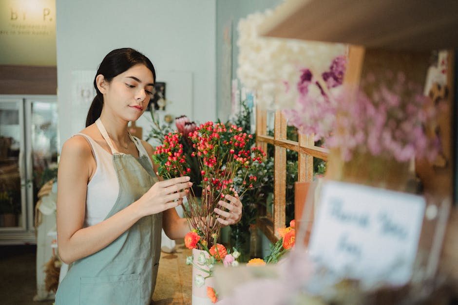 A young female florist with dark hair, wearing a beige apron over a white top, stands in a well-lit flower shop. She is carefully arranging a bouquet of small, vibrant pink and red hypericum berries w