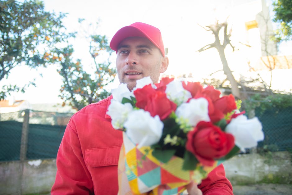 A male delivery person dressed in a red uniform and matching cap is holding a vibrant bouquet of fresh flowers, featuring red and white roses, with green foliage. The bouquet is wrapped in colorful, p