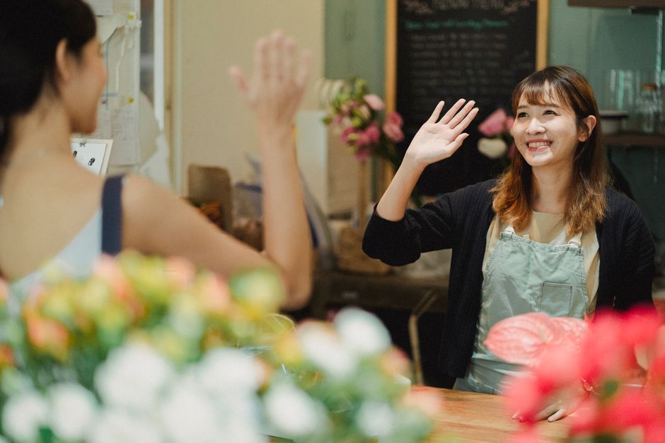 A female florist with shoulder-length brown hair, smiling and wearing a light-colored apron over a beige top, is engaging in a friendly high-five with a customer inside a flower shop. The customer, vi