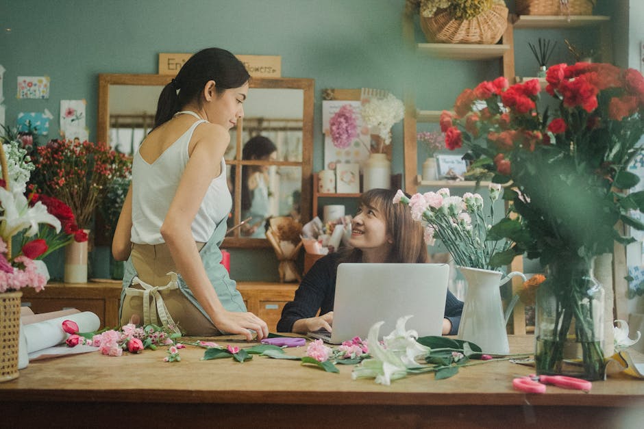 A florist and a customer inside a floral shop, engaging in a conversation about flower arrangements. The florist, wearing a light-colored apron, stands beside a wooden table covered with various flowe