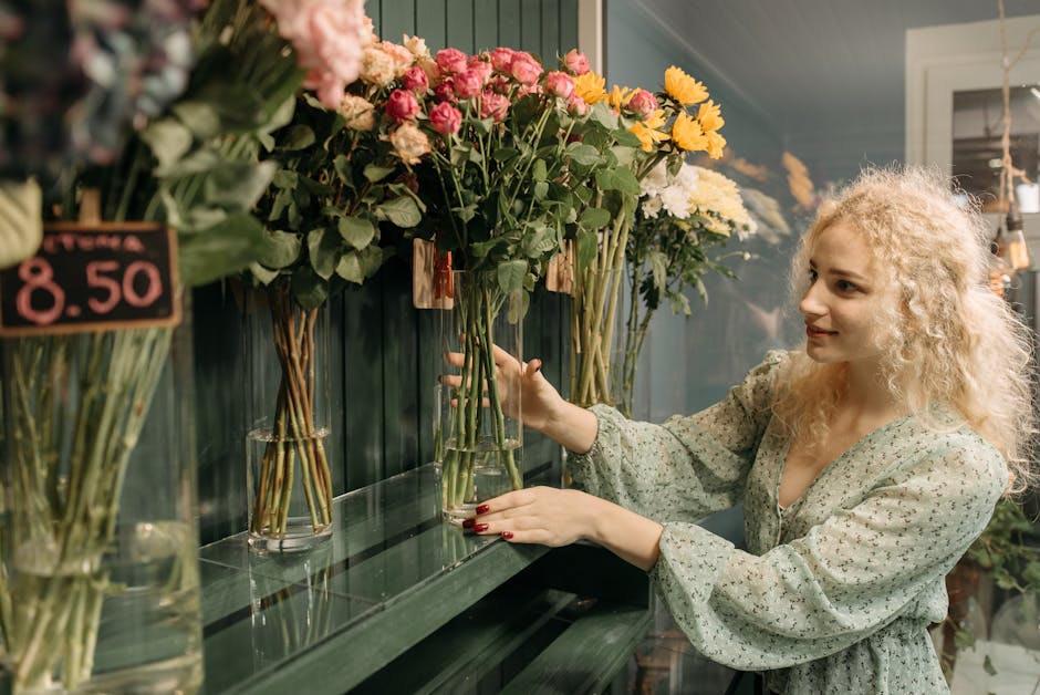 A woman with curly blonde hair selecting a bouquet of fresh pink and yellow flowers in a florist shop. She is holding a glass vase with a bouquet containing pink roses and yellow chrysanthemums. The a
