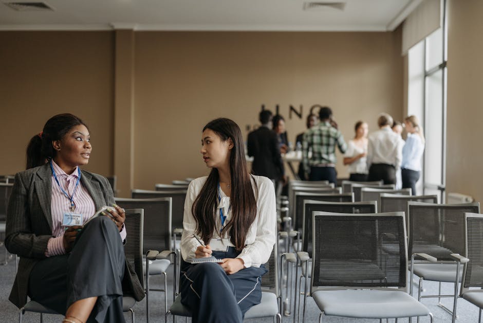 Two professional women seated in a modern conference room engaged in conversation; one is an African British woman with a black blazer and striped blouse, holding a notepad, while the other is an Asia