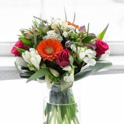 In this elegant Lambeth bouquet, a tapestry of warm, inviting hues meets remarkable structure and nuance, crafted with evident care by local florists inspired by the neighbourhood's rich gardens. Creamy, soft-toned gerberas create a gentle ring, their petals wrapping around brilliant orange hearts that radiate like lanterns lending glow to a Kennington terrace. Velvety magenta roses offer a counterpoint, their deep saturation hinting at love stories unfolding along the Thames. Soft ivory lilies and airy sprays of green contribute luminous freshness and a natural garden feel. Generous, paddle-shaped aspidistra leaves curve protectively at the bouquet's base, enclosing the stems and grounding the more effervescent colours in lush, organic strength. Between the larger blooms, smaller pointed leaves and glimpses of delicate pink filler flowers create a dance of texture and subtle movement, echoing the shifting cityscape outside. Light gently caresses each petal, amplifying vibrancy and making every colour sing. The whole ensemble rests in a slender glass vase, exposing intertwined stems that symbolize connection and unity-perfect for gatherings, celebrations, or heartfelt gifts marking new beginnings in Lambeth.